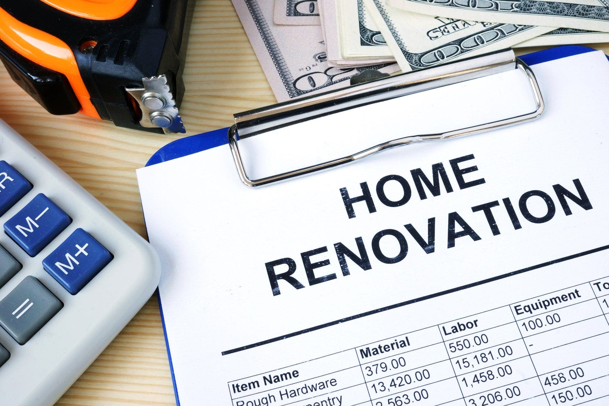 A clipboard holds a home renovation budget sheet and sits next to a calculator, tape measurer, and some cash.