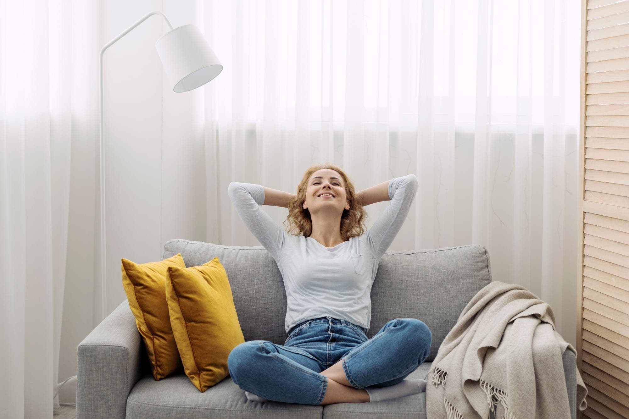 A smiling person relaxing back on their decorated couch, holding their hands behind their head in content.